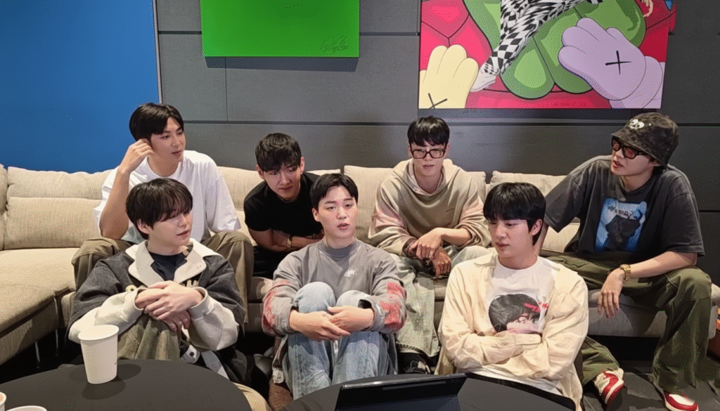 Seven members of a K-pop group sit together on a couch in a studio setting, appearing relaxed while talking during a casual group discussion.