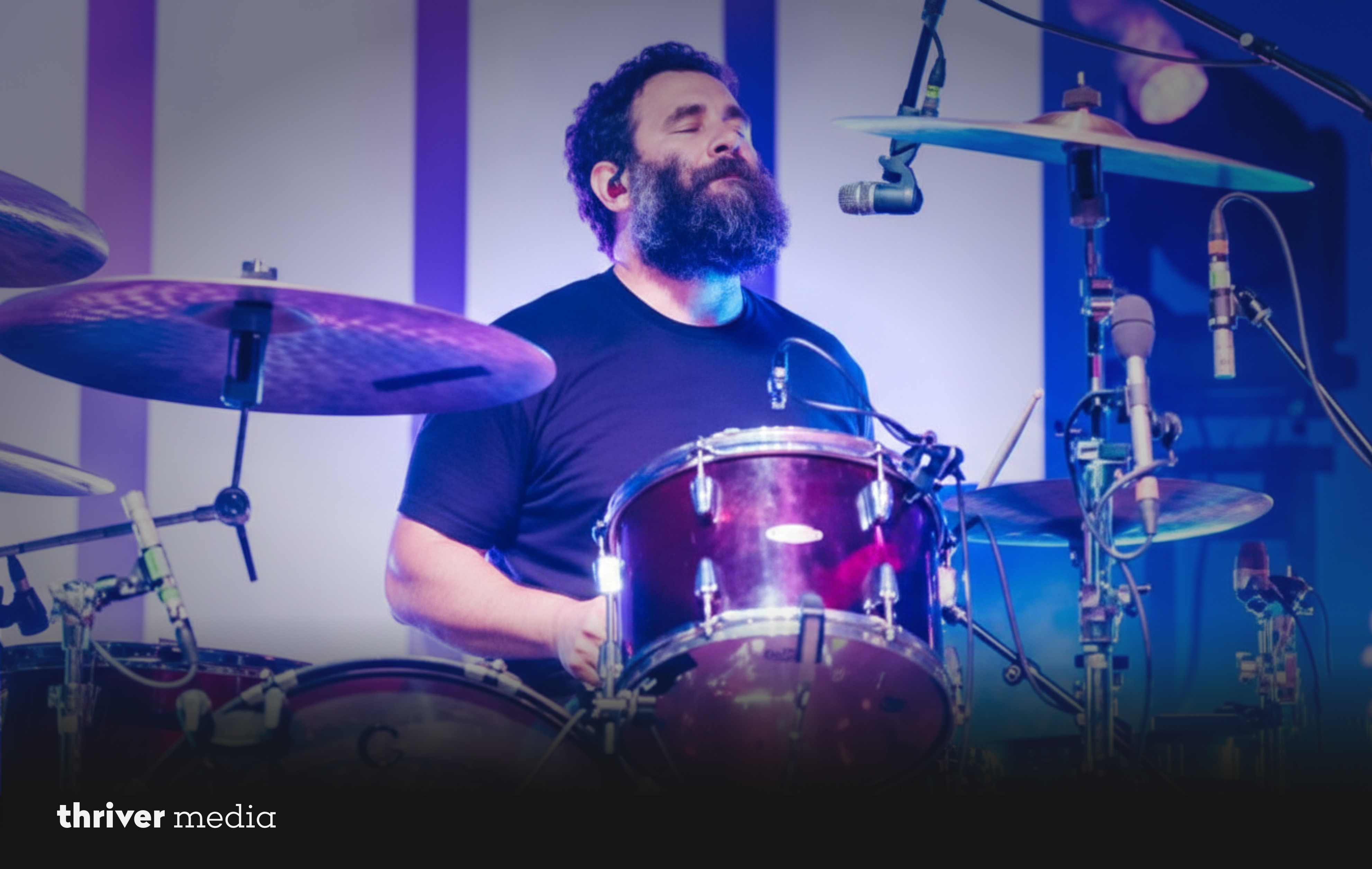 Timothy Very performing live on stage behind a drum kit with eyes closed under blue and purple stage lighting.