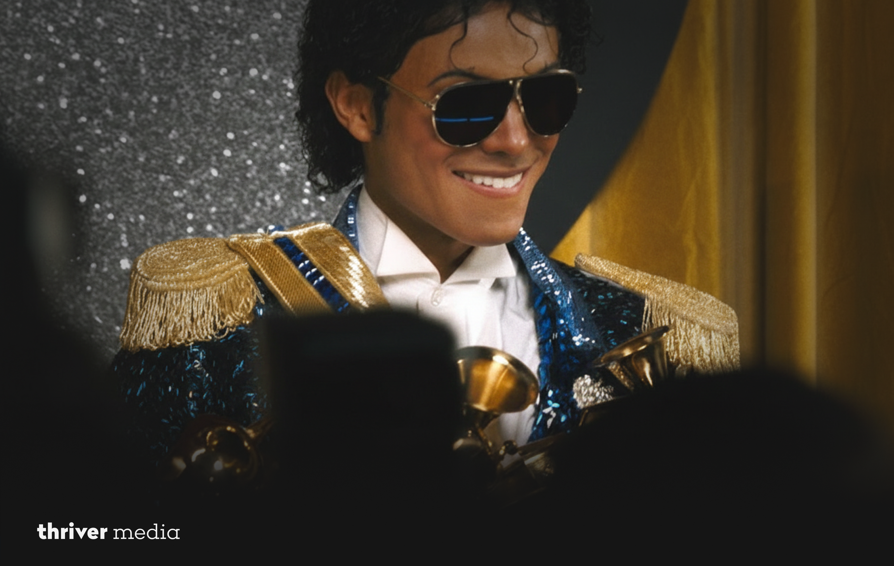 Close-up of Michael Jackson wearing aviator sunglasses and a blue sequined military-style jacket with gold epaulettes, smiling while holding multiple gold awards at a formal event.
