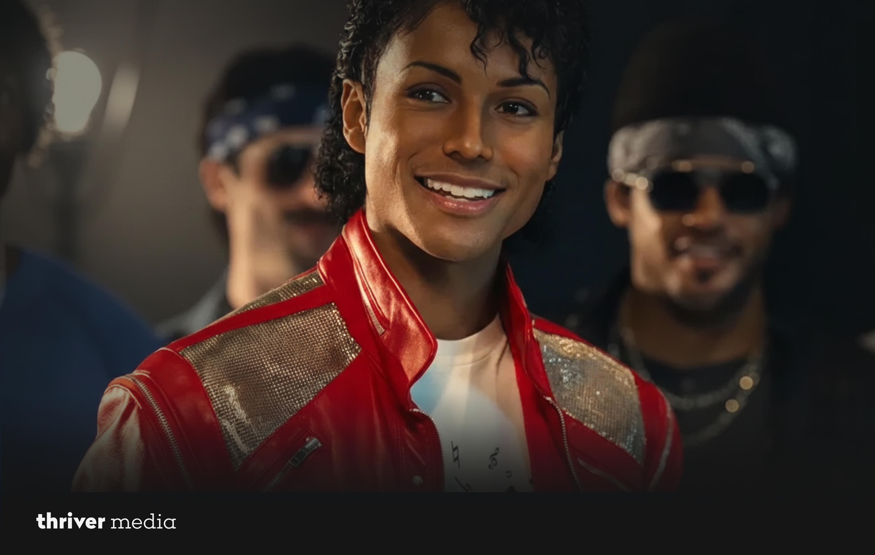 Michael Jackson wearing a red leather jacket with metallic detailing, smiling warmly while standing in front of backup dancers in a dimly lit setting.