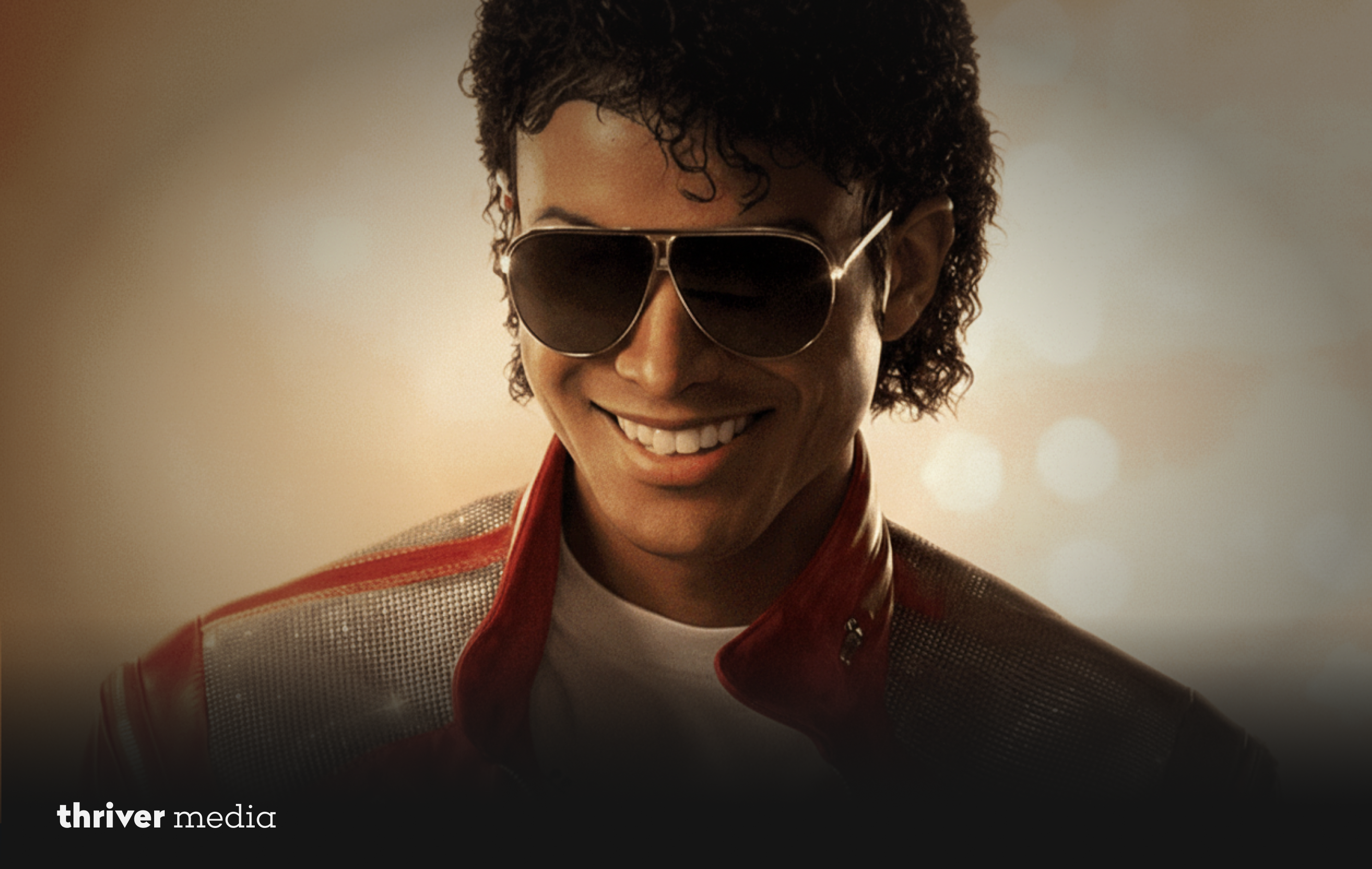 Close-up of Michael Jackson wearing aviator sunglasses and a red jacket with metallic detailing, smiling against a softly lit background.