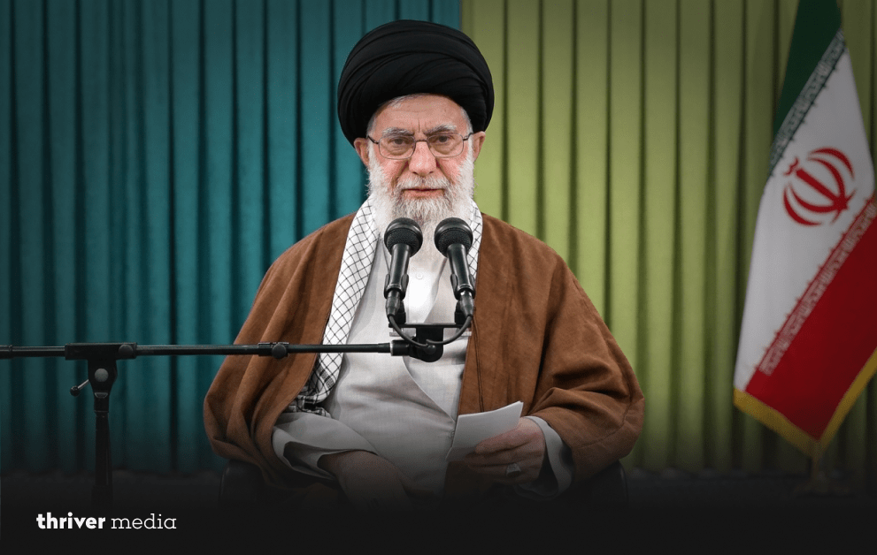 Ayatollah Ali Khamenei (1939-2026): History Bows Its Head 3 Ayatollah Ali Khamenei seated and speaking into two microphones, holding a paper, with green drapes and the Iranian flag visible in the background.