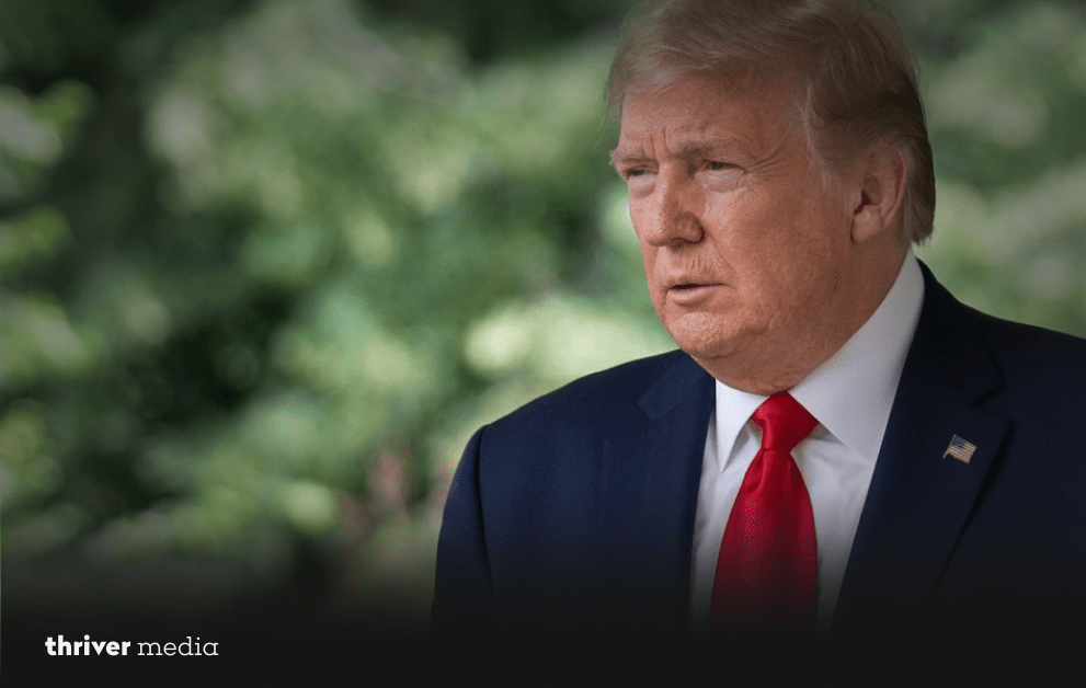 Donald Trump in a dark suit and red tie speaking outdoors with a blurred green background.